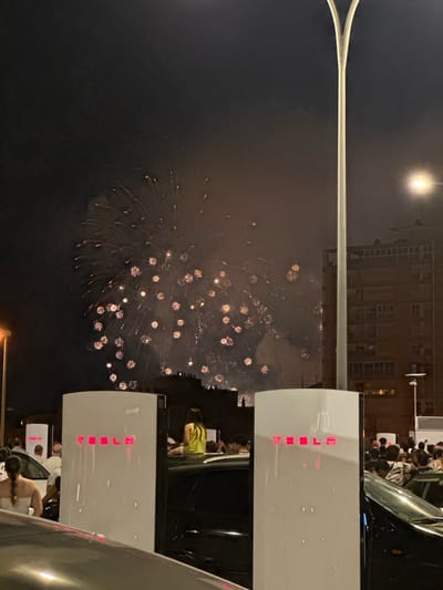 The image captures a nighttime scene in Alicante, Spain, likely during a festive event, as evidenced by fireworks illuminating the sky. In the foreground, two white Tesla Supercharger stations are prominently displayed, their red "TESLA" logos glowing. Several cars, including a black vehicle partially obscuring the right Supercharger, are parked around the charging stations.

A crowd of people is visible, some standing near the chargers and others observing the fireworks. A woman in a yellow top is seated on the roof of a car, facing away from the camera and towards the fireworks. Another woman with braided hair, wearing a white strapless top, is positioned in the lower left of the frame, facing away from the camera.

In the background, a tall, modern building with numerous windows stands silhouetted against the dark sky. The fireworks are a significant element, with numerous bursts of light and smoke spread across the upper half of the image. A tall, curved light pole with a single light fixture is positioned to the right of the frame, extending upwards. The overall atmosphere suggests a public gathering enjoying a fireworks display while in proximity to electric vehicle charging infrastructure.