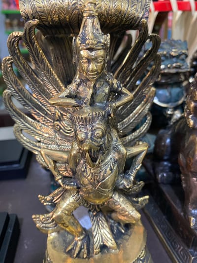 The image depicts a close-up view of a golden, intricately detailed statue of a dragon-like creature with feathers, located in Nusa Dua, Indonesia. The statue appears to be made of metal, possibly brass or bronze, and is positioned at an angle, capturing the head and upper body of the creature. The surrounding environment is blurred, suggesting an indoor setting.