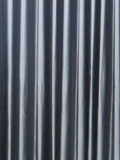 The image displays a vertically oriented, full-frame view of a textured surface, characterized by a series of parallel, vertical ridges and troughs. This creates an alternating pattern of bright, reflective highlights and darker shadowed areas, consistent with corrugated metal or ribbed fabric. The color palette is monochromatic, primarily grey tones with silvery accents. No individuals, discernible objects other than the textured material, or specific actions are visible within the frame. The visual content provides no information to identify a specific location, including any location within the city of Thành phố Đà Lạt, Vietnam.
