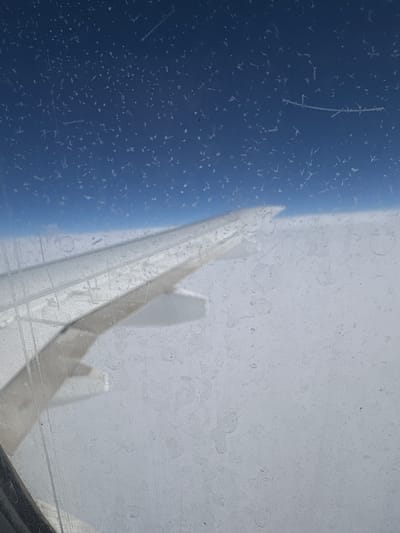 The image shows a view from inside a vehicle, possibly an airplane, looking out through a dirty window. The left side of the window reveals a blue sky with what appears to be frost or condensation forming intricate patterns. The right side of the window is obscured by dirt and grime, making it difficult to see what is outside. There's a blurred shape of what seems to be an airplane wing or part of the vehicle's structure visible through the dirty portion of the window. The location is unknown. There is no indication of the location being in Bratskoye, Russia.