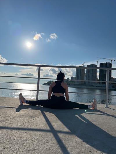 The image depicts a woman performing a split on a concrete platform overlooking a body of water. She is facing away from the camera, wearing a black sports bra and black leggings, with orange and white athletic shoes. The sun is shining brightly in the sky, casting long shadows on the ground. In the background, several tall buildings and construction cranes are visible, suggesting an urban environment. The body of water appears to be a river or bay, with sunlight glinting off its surface. The scene takes place on a clear day with a few scattered clouds. The location is the embankment in Khabarovsk, Russia, along the Amur River, near the construction site of the Khabarovsk Arena.