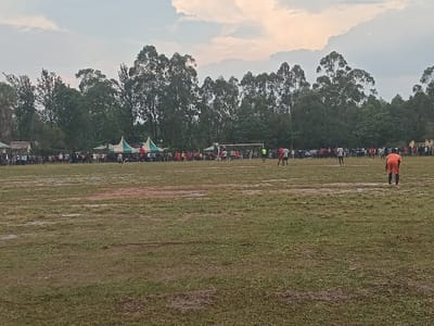 A soccer game is in progress on a muddy field, with players visible scattered around the pitch. A goal is centered in the distance. Spectators line the field, and tents are set up along the edge. Trees form the backdrop under a cloudy sky.