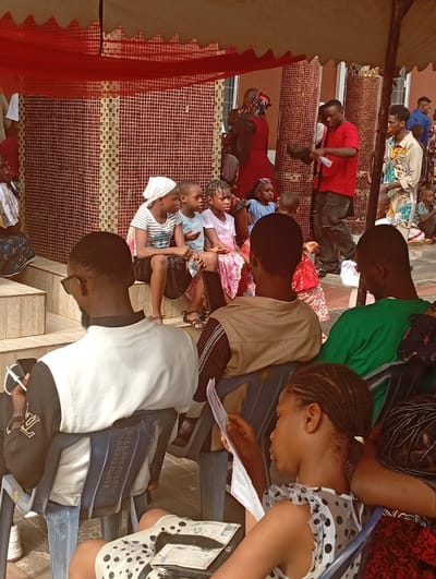 An outdoor gathering shows several children seated on a raised area under an awning. A person in red is talking nearby. Adults seated in chairs face the children. A woman in a polka dot dress is reading something. Other adults stand or sit in the background. Most of the people are of African descent. The scene appears to be a casual event or presentation.