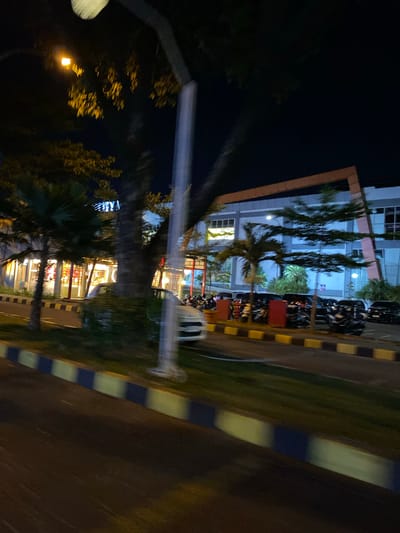 This media file depicts a night scene in Madiun, Indonesia, likely on a street adjacent to a modern building. The image is oriented vertically, suggesting it was taken from a moving vehicle or at an angle.

The primary subjects are a multi-story building with a light-colored facade and orange accents, reflecting the lights from within. Several rows of motorcycles are parked in front of the building, indicating a public or commercial space. Tall, slender trees with dark foliage stand in front of the building, their forms blurred by the low light and motion. Palm trees are also visible, their fronds illuminated by the building's interior lights.

The scene appears to be set at night, evidenced by the dark sky and the illuminated windows of the building. The lighting is artificial, coming from streetlights, the building's interior, and the parked motorcycles' lights. There are no discernible weather conditions, but the reflections on the ground suggest a possibly damp or smooth surface.

Notable details include a striped curb marking a boundary between the road and the green area in front of the building, featuring alternating yellow and dark blue or black stripes. A blurred white pole, possibly a lamppost, is visible in the foreground on the left. The building's windows reveal bright interiors with what appears to be shelving or displays, hinting at shops or offices. No specific activities or events are clearly depicted, but the presence of parked motorcycles suggests it's a location frequented by people. No text is visible in the image. The overall mood is quiet and urban, typical of a commercial area at night.