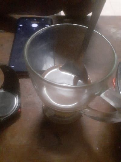 A top-down view depicts a clear glass mug containing a dark liquid and a spoon on a wooden surface in Cimenyan, Indonesia. To the left of the mug, a dark smartphone with its screen illuminated, displaying a camera application, rests horizontally. Further to the left, a portion of a smartwatch or similar wrist-worn device, featuring a metallic rear and a small red illuminated indicator, is partially visible. The wooden surface exhibits a polished or lacquered finish, reflecting light sources. The background is dark and out of focus, suggesting an indoor setting with ambient lighting. No individuals are visible.
