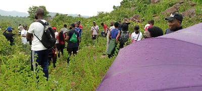The image depicts approximately 20-25 individuals, predominantly young adults of African descent, situated on a narrow, unpaved path traversing a densely vegetated, hilly landscape in Girei, Nigeria. The foreground on the lower right is partially occupied by a wet, purple umbrella displaying visible water droplets. The individuals in the midground are dressed in casual outdoor attire, including t-shirts, pants, and several with backpacks and caps. They are spread out along the path amidst lush green foliage, primarily low-lying shrubs. Actions include standing, conversing, and one individual in the mid-right background holding a mobile phone upwards, suggesting photography or recording. The background features ascending terrain with visible rocky outcrops and a more heavily forested hillside. Distant hills are discernible under an overcast sky.