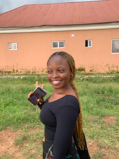 A young woman with braided blonde hair, wearing a black long-sleeved shirt and a patterned skirt, is pictured from the chest up, holding a smartphone in her right hand and making a peace sign with her left. She is smiling and looking to her left, with her head tilted slightly. Behind her, a light orange building with a red tiled roof stands under a cloudy sky. The building has several rectangular windows. The area in front of the building is covered in green grass and some dirt patches. The scene appears to be outdoors, possibly in a residential area in Gwafan, Nigeria.