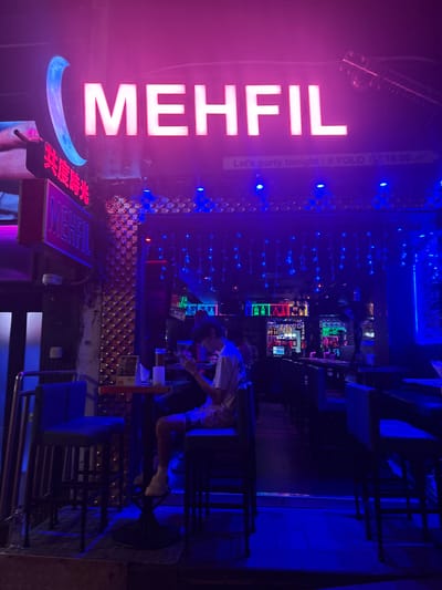 A vertical, medium-shot photograph shows the front and part of the interior of "MEHFIL", a bar in Hong Kong, China.

The bar's facade displays large vertical neon signs with the name "MEHFIL" in white letters and a smaller sign in Chinese characters. A sign says “Let’s party tonight! # Yolo @ 19.00”. Blue lights adorn the entrance. The bar's interior is dimly lit with blue and red lights, revealing bar stools, tables, and bottles of alcohol on the shelves. A person is seated at the bar, facing away from the camera. They are dressed in light-colored attire. Their head is bent, with their focus directed downward.

The foreground shows stacked bar stools with blue seats and metal frames, resting on the ground outside the bar. The floor is a weathered, gray material. The background consists of a wall decorated with metallic tiles reflecting the colorful lights.