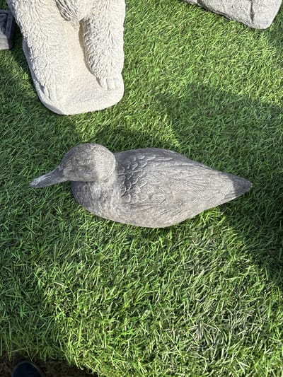 The image shows a gray concrete bird sculpture lying on green artificial turf. The sculpture depicts a bird with its head turned down and to the left. Another concrete sculpture of an animal rests partially visible on the left edge of the frame. A small portion of a dark shoe is visible in the bottom right corner. The scene is outdoors and well-lit. Based on information available, this image was taken in Chirk, United Kingdom.