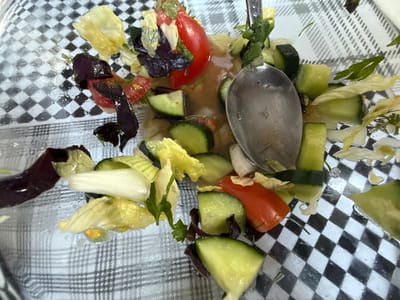 This is a close-up, overhead shot of a salad in a clear bowl. The salad is composed of chopped cucumbers, cherry tomatoes, lettuce leaves (including red and green varieties), and what appears to be a light dressing. A silver spoon is positioned in the center of the bowl, partially submerged in the salad and dressing, suggesting that someone was in the process of eating or serving it. The salad is placed on a surface with a black and white checkered pattern, possibly a tablecloth or placemat. The lighting suggests it is daytime, and there are no external environmental cues. The image is a still life of food, with no people or animals visible. The setting appears to be indoors, likely a dining area. No text is visible in the image.