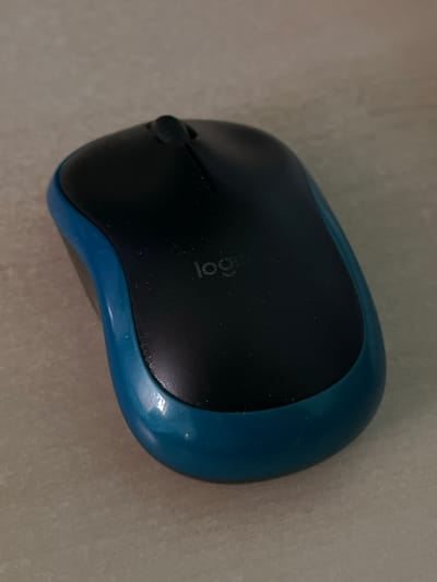 A blue and black Logitech wireless mouse is positioned on a speckled, light-colored surface, resembling a tiled floor. The mouse is angled, with its front facing slightly to the left. The top surface of the mouse is black, featuring the Logitech logo in white. A thin seam runs down the center of the top surface, indicating the separation between the left and right click buttons. A small, dark scroll wheel is visible at the top of the seam. The sides and underside of the mouse are a vibrant blue. The lighting in the image is muted, casting a soft shadow to the right of the mouse. The surface it rests on has a subtle, mottled texture with visible grout lines between the tiles. The specific location of this scene in Lviv, Ukraine cannot be determined from the provided image.