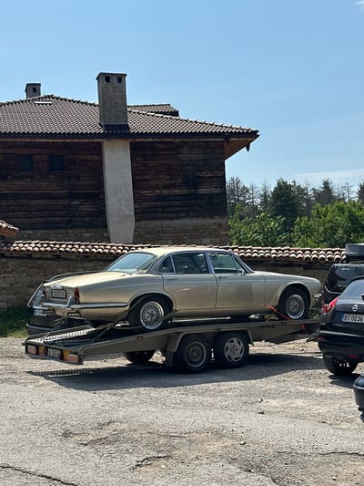 In Arbanassi, Bulgaria, on a sunny day, a vintage gold Jaguar is transported on a car carrier trailer. The car is positioned lengthwise on the trailer, with its rear end slightly elevated. Behind the Jaguar, a black Nissan is parked. The backdrop features a traditional Bulgarian house with a wooden facade and a tiled roof, complemented by a stone wall and lush green trees. The ground is a rough, grey gravel surface. The scene appears to be a transportation or storage area.