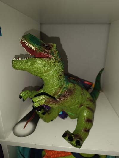 The primary subject of the image is a large, green plastic toy dinosaur, likely depicting a Tyrannosaurus Rex or a similar theropod. The dinosaur has a textured, scaly body with various shades of green, accented by darker brown stripes and patches, particularly on its back and legs. Its mouth is wide open, revealing numerous white, pointed teeth and a bright red interior. The toy is posed in an upright position, standing on its robust hind legs with its smaller front limbs raised. Its eyes appear dark.
The scene is set within a simple, white shelving unit, which provides a clean, neutral background. The walls behind the dinosaur are light-colored, forming a corner, and a prominent shadow is cast on the wall, indicating an indoor setting with a light source positioned to the front or side.
The dinosaur toy is static, captured in a posed stance that suggests roaring or an active moment, though it is an inanimate object.
Notable details include a glimpse of a colorful, blurred object (possibly another toy or item) with hues of purple, red, and blue, situated behind the dinosaur's mid-section. Below the shelf on which the dinosaur stands, a portion of an orange and blue object, possibly fabric, is visible in the lower section of the frame. An elongated, dark object with a reddish-brown tip lies on the white shelf surface near the dinosaur's left front limb. The overall lighting is somewhat dim, contributing to the definition of shadows.
No visible text can be deciphered on the dinosaur or within its immediate surroundings. While the image is contextualized as Simferopol, Ukraine, no specific visual cues within the photo itself confirm this location.