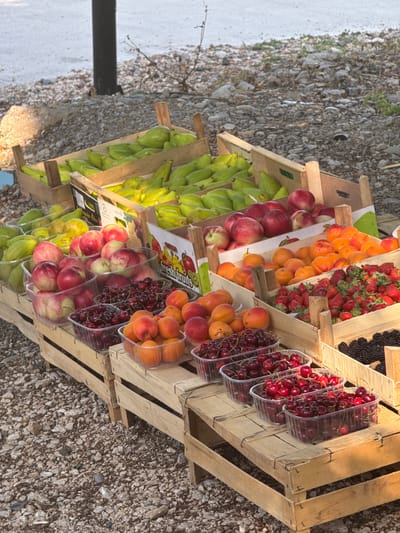 This image captures a vibrant outdoor fruit stand in Kamenovo, Montenegro. In the foreground, several wooden crates are stacked, displaying an array of fresh fruits. The crates hold plastic containers filled with cherries, apricots, and strawberries. Behind these, larger wooden crates overflow with apples and a variety of green, oblong fruits, possibly pears or unripe bananas. Further back, more crates of these green fruits are visible. The ground surrounding the fruit stand is covered in gravel and small stones. A dark, vertical pole, likely a street light or signpost, stands in the background, partially obscuring the view of the sea, which appears as a textured, light-colored expanse. The lighting suggests it is daytime, with shadows cast by the fruit stand and the pole. There are no people visible in the frame, and the focus is solely on the abundant fruit display.