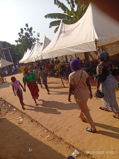 The image documents an outdoor scene in Isinyi-Nando, Nigeria, at 15:52 on December 19, 2025. The ground is unpaved, reddish-brown earth, uneven, and contains scattered debris. A shallow, linear depression is visible across the foreground. Bright daylight casts distinct shadows.
In the mid-ground, a row of five or more white, peaked event tents (canopies) is erected. One tent prominently displays partial red lettering, including "ENTERIA BLESSING." Beneath these tents, numerous individuals are gathered, some seated on chairs, suggesting an event is in progress. The background features green foliage, including palm trees, a metal water tower structure on the far left, and a brick wall section on the right.
In the foreground and mid-ground, five individuals are in motion. On the right, an individual with bright purple hair, wearing a beige long-sleeved top and matching trousers, walks away from the camera. Immediately to their right, another individual, seen from behind, wears a dark patterned dress, a dark headwrap, and carries a dark handbag; this person is also walking away. On the left side of the frame, an adult in a green top and red patterned skirt holds hands with a child in a pink top and dark shorts; both walk towards the left. Another indistinct individual is visible walking further back on the left side.