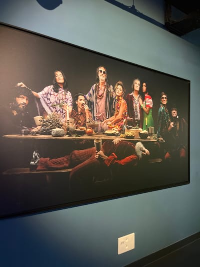 A large, rectangular, black-framed photographic print is displayed on a solid blue-green wall within The Consortium Museum (Le Consortium) in Dijon, France. The print depicts a modern reinterpretation of "The Last Supper," featuring ten individuals gathered around a long, rustic wooden table against a dark, featureless background.
Nine individuals are seated upright behind the table, arranged in a line. Their attire is varied, generally reflecting bohemian or hippie-style aesthetics with flowing fabrics, patterned garments, necklaces, and sunglasses.
From left to right:
1. A man with dark, curly hair and a beard, wearing dark clothing. His left hand is near his head.
2. A woman with long dark hair, wearing a purple and white patterned kaftan-like garment and holding a small plant branch.
3. A man with dark hair and a mustache, wearing a patterned shirt, looking down and eating from a bowl.
4. A man with long dreadlocks, a beard, and sunglasses, wearing multiple necklaces and a light-colored, patterned shirt under a robe.
5. A woman with curly hair, wearing a vibrant multi-colored patterned dress and large earrings, looking to her right.
6. A man with graying hair, wearing multiple necklaces and a reddish-brown patterned jacket, looking upwards.
7. A woman with dark hair, wearing a red and white striped top and possibly green pants, holding a lit candle.
8. A man with sunglasses, wearing a patterned green and blue jacket or shirt.
9. A man with dark hair, wearing a dark robe or jacket.
In the foreground, an older man with a long white beard is lying horizontally on a wooden bench in front of the table. He wears a red velvet-like jacket and reddish-brown patterned pants, holding a brass-colored goblet in his right hand and looking towards the viewer.
The wooden table is laden with various objects, including ceramic bowls, plates, clusters of dried herbs or flowers, candles in holders, a round cheese-like object, and an assortment of fruits such as grapes and apples.
The wall on which the print hangs is a uniform blue-green color. Reflections from an overhead light fixture are visible on the upper part of the print. A white electrical outlet with two sockets is present on the wall below the print, positioned to the right. The floor appears dark.