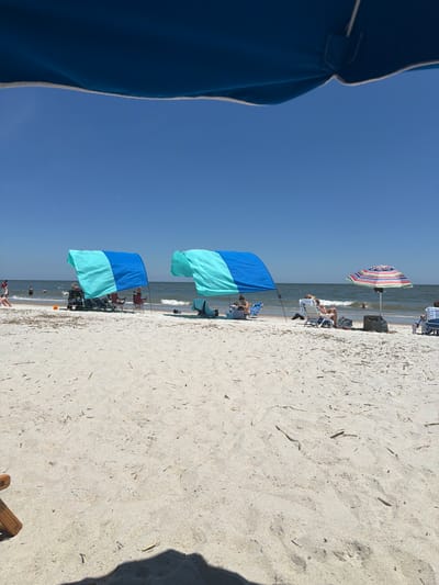 The image depicts a sunny day at a sandy beach on Hilton Head Island, United States of America. The foreground is dominated by sand, with scattered pieces of dried seaweed or driftwood. A portion of a blue beach umbrella is visible at the top of the frame, casting a shadow on the sand. In the middle ground, two blue and turquoise beach tents are set up, providing shade for several people seated in beach chairs. To the right of the tents, a colorful striped beach umbrella is erected, also offering shade to individuals in chairs. In the background, the ocean stretches to the horizon under a clear blue sky, with gentle waves breaking on the shore. Several people are visible in the water and along the shoreline, enjoying the beach.
