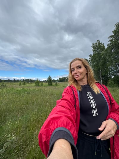 A woman with blonde hair smiles at the camera while wearing a red jacket over a black t-shirt. The t-shirt has white text in a rectangular box that reads "DANCE WORLD TOUR." She is outdoors in a grassy field with trees in the background under a cloudy sky. The image appears to be taken during the daytime. The scene suggests a casual outing or a moment of enjoyment in nature. The location context is Mars, Russia.