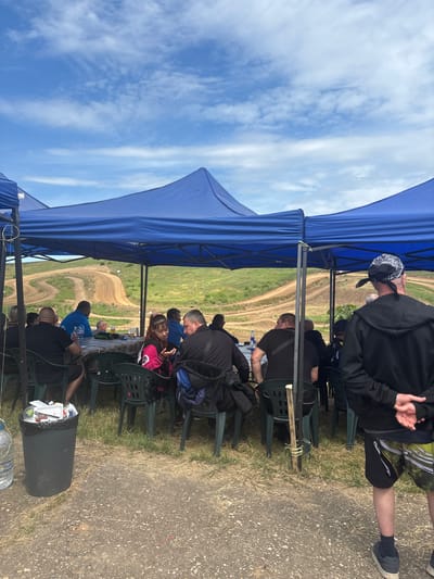A group of people are seated outdoors under a blue canopy, likely watching an event. The location appears to be a dirt track or field, with a visible dirt road running in the distance. The sky is blue with scattered clouds. A trash can filled with refuse is visible near the seating area. A man stands at the edge of the canopy, while others are seated on green chairs at a table. The scene appears to be in Kalipetrovo, Bulgaria.