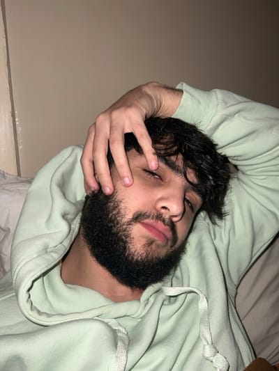 A man with dark hair and a beard is lying down, wearing a light green hoodie. His head is tilted, and he is resting his left hand on his face. He appears to be relaxed or tired, with his eyes partially closed. The background is a blurred, light-colored surface, possibly a wall or bedding.
