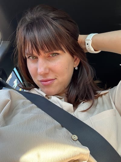 A selfie shows a fair-skinned woman with light brown hair and bangs. She has green eyes and is wearing a seatbelt. Her right arm is visible, with a white watch on her wrist. She wears a small, square earring. The background appears to be the interior of a car, with a glimpse of an exterior structure visible through the car window. The location cannot be determined.