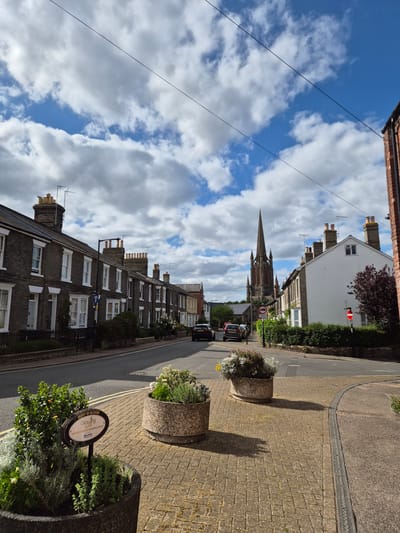The image depicts a street scene in Bury St Edmunds, West Suffolk, United Kingdom. On the left, a row of terraced houses with dark brick facades and white-framed windows stretches along the street. Several of these houses have chimneys and aerials on their roofs. A black car is parked on the street in front of the houses, and another black car is driving away from the camera.

In the center of the image, a tall, spired church building dominates the skyline, with intricate architectural details. Adjacent to the church, on the right side of the street, is a white building with a red-tiled roof. A dark red tree with dense foliage stands next to this building.

The street is paved with asphalt, and the sidewalk is made of yellow and grey brick pavers. Two large, circular concrete planters containing greenery and flowers are positioned on the sidewalk in the foreground. One planter has a sign attached to it that reads "BURY IN BLOOM Growing well NEIGHBOURHOOD PLANTING SPACE Suffolk".

The sky is partly cloudy, with a mix of white and grey clouds against a bright blue background. Power lines are visible crisscrossing the sky. Several traffic signs are present on the street, including a blue directional sign and two red circular signs with white bars, indicating prohibitions. The overall atmosphere is that of a residential street on a bright, possibly sunny day.