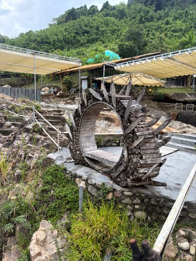 The image shows a rustic outdoor seating area with a distinctive circular wooden structure in the foreground. This structure, made of stacked logs and branches with pointed wooden extensions, resembles a wheel or a natural archway and has a wooden bench inside. The area appears to be a resort or park, set against a lush, green mountainside. In the background, there are several canvas awnings providing shade, some tables, and chairs, suggesting a place for relaxation. Stone steps and rocky terrain are visible, indicating a natural, uneven landscape. Two people are partially visible in the mid-ground, near the steps, interacting with the environment. The overall scene is outdoors, with vegetation and natural rock formations dominating the landscape.