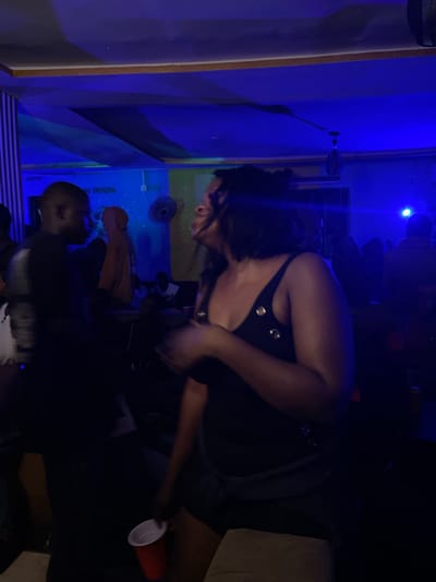 In a dimly lit interior, likely a bar or club in Unknown, Nigeria, a woman with dark hair is captured mid-motion, her head tilted back as if laughing or singing, her mouth open. She wears a black tank top with two silver buttons. To her left, a dark-skinned man, seen from the side, is silhouetted against the blue-tinged lighting. He appears to be wearing a black t-shirt. In the background, other patrons are visible, some seated, creating a sense of a social gathering. The environment is characterized by blue and some yellow lighting, with what appears to be a "NO SMOKING" sign and a fan mounted on the wall. A red plastic cup is held in the foreground by an unseen person. The overall scene suggests a lively atmosphere within the establishment.