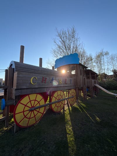The image displays an outdoor playground scene in Niederanven, Luxembourg, under a clear, bright blue sky. The central subject is a large wooden play structure designed to resemble a train, complete with a front boiler section and multiple carriage-like segments. No people are present, giving the scene a serene atmosphere.
The wooden structure is detailed with six prominent, decorative yellow wheels featuring red spoke patterns, three on each side. A distinctive blue, wave-shaped element is attached to the front of the train structure. Along the side of the train, the name "CHARLY" is spelled out in individual, colorful block letters: a yellow 'C', orange 'H', blue 'A', red 'R', yellow 'L', and orange 'Y'.
In the background, trees with mostly bare branches suggest an early spring or late autumn setting. A silver slide is visible on the far side of the wooden structure, alongside various ladders and platforms, confirming its use as a children's play area. The sun is low in the sky, creating a strong lens flare that partially brightens the train's side and casts long shadows across the neatly mown green grass, indicating either early morning or late afternoon. The weather is sunny and cloudless.