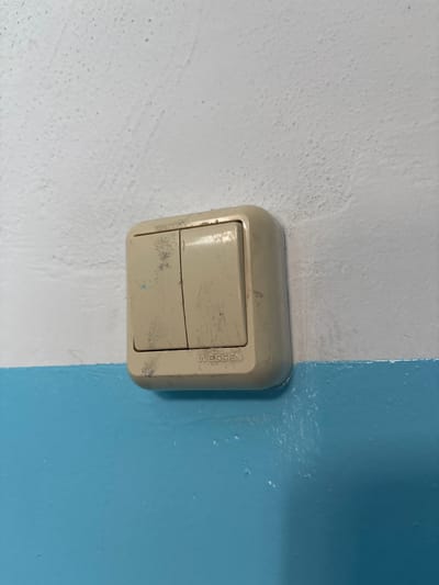 The image shows a close-up of a beige, two-gang light switch with the brand name "WESSEN" visible. The switch is mounted on a wall that is painted white on the top half and a bright blue on the bottom half. The white portion of the wall has a textured, slightly speckled appearance, while the blue portion is smoother and appears to be a glossier finish. The light switch itself appears worn, with scuff marks and some blue paint transfer on its surface, suggesting it may have been exposed to painting work or general wear and tear. There are no people or any other objects clearly visible in the frame, and the image focuses solely on the light switch and the wall it is mounted on. The scene suggests an interior setting, likely a domestic or commercial space. No specific location in Blagoveshchensk, Russia can be identified from this image alone.