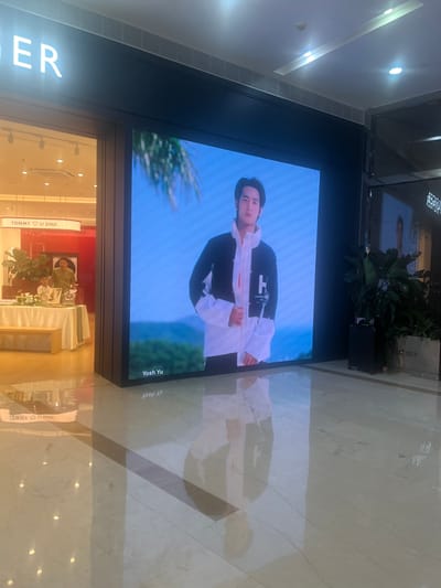 In the Liandu District of China, a digital billboard displays a young man wearing a black and white jacket with the letter 'H' prominently featured. The billboard is situated within a retail environment, indicated by a store entrance to the left with a sign that reads "TOMMY ♡ LI SHUI." Inside the store, two individuals are visible near a table adorned with flowers and other display items. The floor reflects the billboard and store lighting, creating a polished and somewhat diffused image. The overall scene appears to be a commercial space within a shopping area.