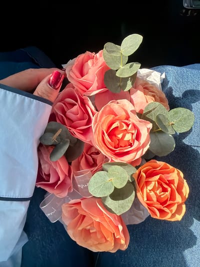 The image displays a close-up, top-down perspective of a floral bouquet observed in Kukui, Russia. The bouquet primarily consists of numerous roses in various shades of peach, pink, and orange, interspersed with several sprigs of round, silvery-green eucalyptus leaves. White tissue paper and a sheer white ribbon featuring faint, illegible cursive script are visible within the arrangement. The bouquet rests upon blue denim fabric, consistent with jeans, on a person's lap. To the left, a light-skinned hand with glossy red fingernail polish is visible, positioned near the flowers. The individual is wearing a white garment with a dark-trimmed cuff. Strong, directional sunlight illuminates the scene, casting distinct shadows across the flowers and denim. The upper-left background is dark and indistinct.