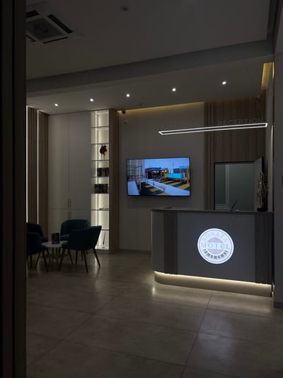 The image captures a modern and well-appointed reception area or lobby, likely belonging to the Sonata Motel in Simferopol, Ukraine. The scene is brightly illuminated by various artificial light sources, suggesting either nighttime or an interior space relying entirely on electric lighting.
At the center-right of the frame, a sleek reception desk features a prominent circular, backlit sign displaying "SONATA MOTEL" with a graphic that resembles piano keys. Above the desk, a vertical strip light runs along a wooden-paneled wall, clearly labeled with the word "RECEPTION." The desk itself has a contemporary design, incorporating light wood accents and subtle backlighting along its base.
The overall decor of the lobby is modern and clean, characterized by white walls and ceilings, complemented by light-colored wooden panels. The floor is covered with large, neutral-toned tiles. Various lighting fixtures contribute to the ambiance, including recessed spotlights in the ceiling and integrated strip lighting along wall panels and shelving units.
On the left wall, a flat-screen television is mounted, displaying an image that appears to be an interior industrial or workshop setting with yellow and black striped objects. Below the television, a long, horizontally oriented shelving unit is built into the wall, showcasing several decorative items and books, also lit by integrated strip lights.
In the foreground, a small waiting area is furnished with two dark teal or green upholstered chairs with wooden legs, arranged around a low, light-colored table. An air conditioning unit is visible on the ceiling in the upper left portion of the image. The area is empty of people, presenting a quiet and inviting atmosphere.