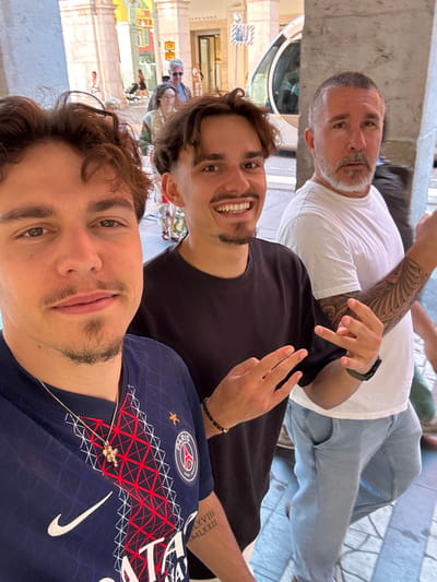 Three men are posing for a selfie outdoors in Nice, France. The man closest to the camera is wearing a Paris Saint-Germain jersey and a silver necklace. The man in the middle is wearing a black t-shirt and making a hand gesture. The man in the back is wearing a white t-shirt, jeans, and a black watch, with a visible arm tattoo. Behind them is a pedestrian street lined with shops, including a Zara Home store. The setting appears to be a sunny day near an arcade or covered walkway.