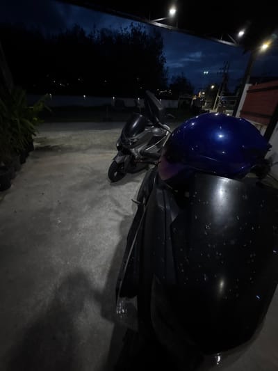 In Wichit, Thailand, at night, two motorcycles are parked on a concrete surface, possibly a driveway or lot. The motorcycle in the foreground is partially visible, with a dark front fairing and a bright blue helmet resting on it. The helmet has a glossy finish and reflects the ambient light. The motorcycle behind it is more fully visible, appearing to be a grey scooter. The background shows a dimly lit street with trees, power lines, and distant lights, suggesting an urban or suburban setting. The sky is dark with some clouds. The scene is lit by artificial lights from overhead fixtures and streetlights.