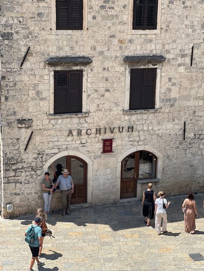 This media file captures a street view in Kotor, Montenegro, bathed in the warm light of what appears to be a sunny afternoon. The scene is dominated by a weathered stone building, its facade made of unevenly shaped blocks. Two dark, louvered windows are visible on the left side of the building, one above the other. To the right, the building features arched doorways and windows. The word "ARCHIVUM" is inscribed vertically in large, dark letters on the stone facade. A small, rectangular plaque is also affixed to the wall below the "M" of "ARCHIVUM."

Several people are present, mostly seen from above as they walk or stand on the cobblestone pavement. On the far right, a woman in a long, light-colored dress walks away from the camera. Further to the left, a group of three people are clustered near an arched doorway. One person wears a white shirt and light-colored pants, another a black dress, and the third, a woman, is in a patterned top and dark skirt. Near them, a man in a checkered shirt and straw hat stands with his arm around a woman in a green shirt and grey pants. Further down the street, another group of people are visible. One individual is wearing a blue backpack and dark shorts, and another has a striped shirt and a brown bag.

The overall impression is of a historical location on a pleasant day, likely a tourist destination given the presence of several casually dressed individuals exploring the area. The sunlight casts distinct shadows on the cobblestones, indicating a clear sky. The architecture, with its stone construction and arched openings, suggests an old European city.