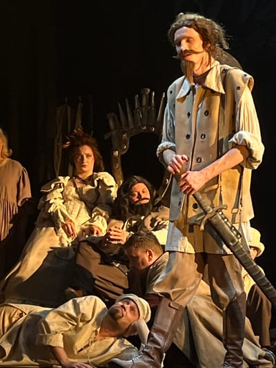 The image depicts a theatrical performance occurring on a stage in a venue located in Novoshakhtinsk, Russia. Six individuals, all adults in period-style costumes, are visible. A tall male figure, characterized by long dark hair, a pronounced handlebar mustache, and a goatee, stands centrally, clad in a light-colored, long-sleeved shirt, a beige waistcoat with dark buttons, and dark knee-high boots. He holds a dark, sheathed sword vertically in his left hand, resting it against his leg, and looks upward and to the right with a serious expression. To his left, a female figure with styled, dark hair and an elaborate light-colored gown with ruffles is seated on a chair, observing forward with a solemn demeanor. Directly behind the seated female, a male figure with dark, curly hair, a full beard, and dark clothing is seated, his hands clasped. To the far left, a partially visible individual in a simple garment is present. In the lower mid-ground, a male is positioned, appearing to be seated or kneeling. In the foreground, another male with a goatee and a light-colored cap is reclining on his back, looking upward. The background is dark, with a noticeable ornate, dark, spiky structure, likely a theatrical prop or part of the set design. Directed stage lighting illuminates the performers.