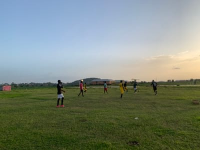 The media file depicts a group of people playing soccer on a grassy field in Gwafan, Nigeria. Several individuals, appearing to be young men, are scattered across the field, some actively engaged in the game, while others are walking or standing. They are dressed in athletic wear, with some wearing brightly colored jerseys. The field is green and appears well-maintained, with some scattered litter. In the background, there are buildings, including what looks like a pink structure with a soccer goal, and a distant tree line. The sky is a clear, pale blue, transitioning to a soft orange and yellow hue towards the horizon, indicating it is either sunrise or sunset. The lighting suggests a peaceful and perhaps cooler part of the day, common for outdoor activities. The overall scene conveys a sense of casual recreation and community engagement in a rural or semi-rural Nigerian setting.