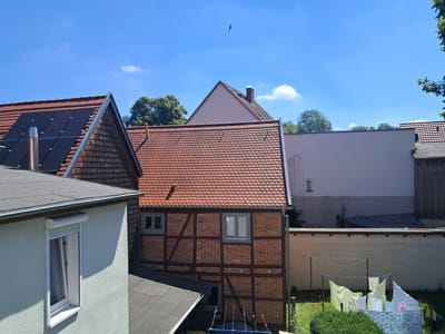 This is an eye-level, outdoor, daytime, medium shot of a residential area in Parchim, Germany. The image captures several houses with different architectural styles and rooftops. In the foreground, a light-colored house with a flat, grey roof and a visible window with a white frame and roller shutter is on the left. Adjacent to it, to the right, is a house with a red-tiled, pitched roof and solar panels on its upper section. Further to the right, a traditional half-timbered house with red brick and dark wooden beams is visible, featuring two windows with grey frames. Behind these structures, a large, pale pink building with a steeply pitched roof and a chimney stands out. Beyond that, a long, light-colored brick wall forms a boundary, with clothes drying on a clothesline in the yard. The sky is clear blue with a few scattered white clouds. The scene suggests a quiet, residential neighborhood on a sunny day.