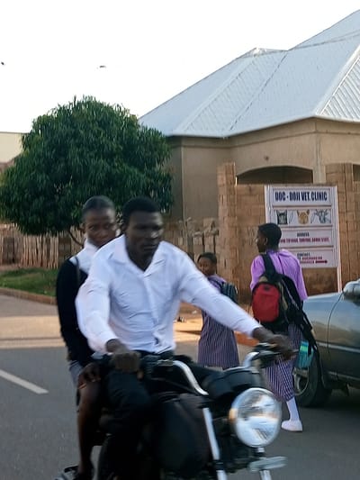 In Tumfure, Nigeria, a man wearing a white collared shirt and dark pants rides a motorcycle, with another person in a black shirt and grey shorts as the passenger. They are on a paved road, with a grassy area and trees to their left. In the background, a building with a light-colored metal roof is visible, along with a sign for "DOC-DOJI VET CLINIC." Several other people are walking in the background, including two wearing purple tops and striped skirts, one with a red backpack. A silver car is parked to the right of the motorcycle. The lighting suggests it is daytime.