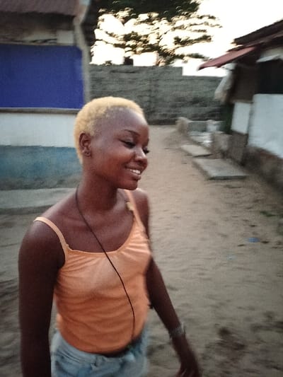 A smiling, dark-skinned woman with short, bleached blond hair wears a pale orange tank top and denim shorts. She stands outside in an area with sandy ground and buildings in the background. A thin black necklace hangs around her neck, and a bracelet adorns her wrist. The lighting suggests late afternoon or early evening.