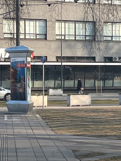 The image depicts an urban outdoor scene in Liepaja, Latvia, dominated by a large, multi-story building in the background. This building features a grey, textured facade with numerous rectangular windows on its upper floors and large, dark-tinted glass panels on the ground floor, where an external air conditioning unit is visible on the right. Bare tree branches partially obscure the building in the upper-mid section.
In the lower-left foreground, a tall, octagonal advertisement column with a grey base and cap displays an advertisement for "Lielais dzintars" (Great Amber Concert Hall), including an image of an individual playing a cello and the text "Liepāja". To the right of this column, a thin metal pole supports a circular blue traffic sign and a smaller red rectangular sign.
An individual, dressed in a dark-colored, possibly long coat or jacket, and light-colored shoes, walks from right to left in the midground, carrying a light-colored bag in their right hand. They are positioned between two rectangular concrete planters, which contain sparse greenery. Other similar planters are arranged in a line leading towards the background, separating a paved sidewalk from a grassy area. The sidewalk, constructed of light-colored rectangular blocks, extends into the foreground with distinct shadows. The adjacent grassy area shows a striped pattern. A silver car's front fender and wheel are partially visible on the far left, behind the advertisement column. The scene is illuminated by strong sunlight, casting visible shadows.