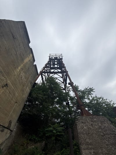 The image shows the abandoned headframe of the Kapan mine in Kapan, Armenia. The headframe is a large, rusty metal structure that is partially obscured by trees. A large concrete wall is visible on the left side of the image, and a large concrete block is in the foreground on the right. The sky is overcast. The scene depicts an industrial ruin overtaken by nature, suggesting a post-industrial landscape within the city of Kapan.