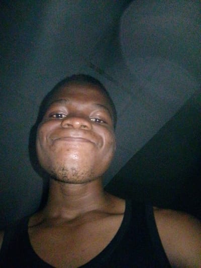 A dark, grainy, close-up selfie of a young Black man is shown. He is wearing a black tank top and looking directly at the camera. His expression is a slight smile or grin, with his lips parted. There are faint dark stubbles on his chin and jawline. The background is dark and out of focus, suggesting an indoor setting with low lighting, possibly a bedroom or dimly lit room in Ipetumodu, Nigeria. The lighting casts shadows on his face, particularly on the right side of his forehead and cheek. The overall impression is that of a casual, unposed snapshot.
