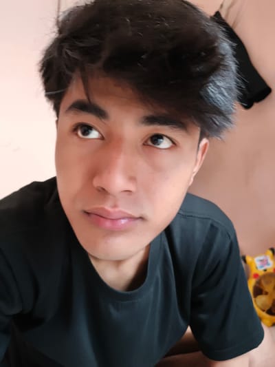 A young man with dark hair and a black t-shirt is looking off to the side. The background is a peach-colored wall. There is a small black garment hanging on the wall and a yellow cartoon container visible in the bottom right corner. The location is uncertain, as there is no readily apparent information to specify the city of Cianjur, Indonesia.