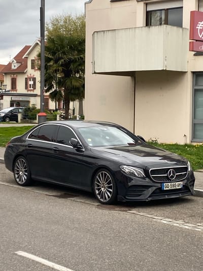 The image shows a black Mercedes-Benz sedan parked on a gray asphalt road in Bischheim, France. The car has a license plate reading "GQ-590-AH." A building is visible behind the car on the left, with a sign on top. In the background, trees and houses are visible, including one with a sign that says "EPICERIE TERAILLON." The sky is overcast.