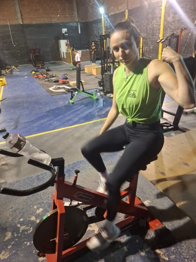 A woman is on an exercise bike in a gym setting. She wears a green T-shirt, black leggings, and white sneakers. She is turned sideways, looking at the camera, and flexing her right bicep. A water bottle is attached to the front of the exercise bike. Various pieces of gym equipment are scattered in the background, including weights, benches, and other exercise machines. The floor is blue with yellow lane markings. Based on the T-shirt, the location is in Wilde, Argentina.