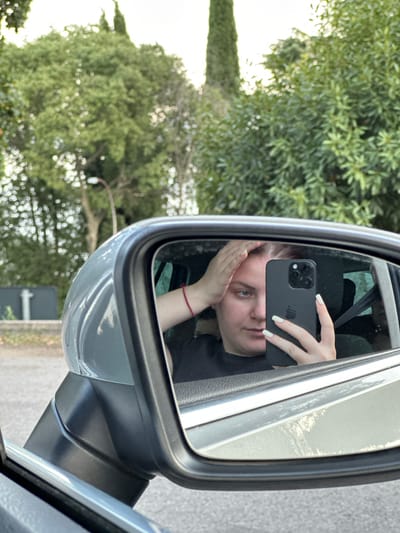 A young woman takes a selfie in the side-view mirror of a car. She is inside the car, seated in the driver's seat, and is looking at her reflection. Her left hand is touching her hair, and her right hand is holding a black smartphone, which is capturing the image. She has on a black shirt and a red bracelet on her left wrist. The interior of the car, including the car's gray exterior, is visible in the mirror's frame. Outside the car, lush green trees and a bright sky are visible. The scene is likely in Lovran, Croatia, given the surrounding vegetation which is characteristic of the region. The setting suggests a daytime outdoor environment, possibly on a street or in a parking area in Lovran, Croatia.
