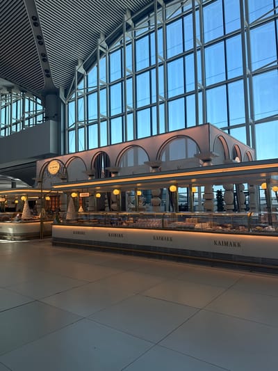 The image depicts the interior of a modern, well-lit establishment, likely an airport terminal in Arnavutköy, Turkey, given the architectural style and the prevalence of large glass windows revealing a bright sky. The primary focus is on a sleek, contemporary food service counter, branded with the name "KAIMAKK" repeated along its facade. The counter features a glass display case filled with various pastries and confections, illuminated by soft, warm lighting. Decorative elements, such as white cone-shaped structures resembling Christmas trees, are also visible.
Above the counter, an arched decorative facade adds a touch of elegance, with subtle lighting accentuating its design. The ceiling is a complex structure of white metal beams and slats, characteristic of modern large-scale public spaces. The floor is tiled in a neutral tone. The overall impression is one of cleanliness, order, and a sophisticated commercial environment, designed to serve customers in a bustling transit hub. There are no people visible in the frame.