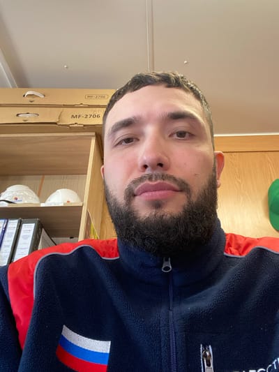 A man with a dark beard and short dark hair is shown in a close-up, head-and-shoulders shot. He appears to be inside a building or workspace, standing in front of shelves with cardboard boxes labeled "MF-2706" and some white hard hats. He is wearing a navy blue fleece jacket with red accents and a patch featuring white, blue, and red stripes reminiscent of the Russian flag. No specific location in Luzhitsy, Russia, can be identified from the image.